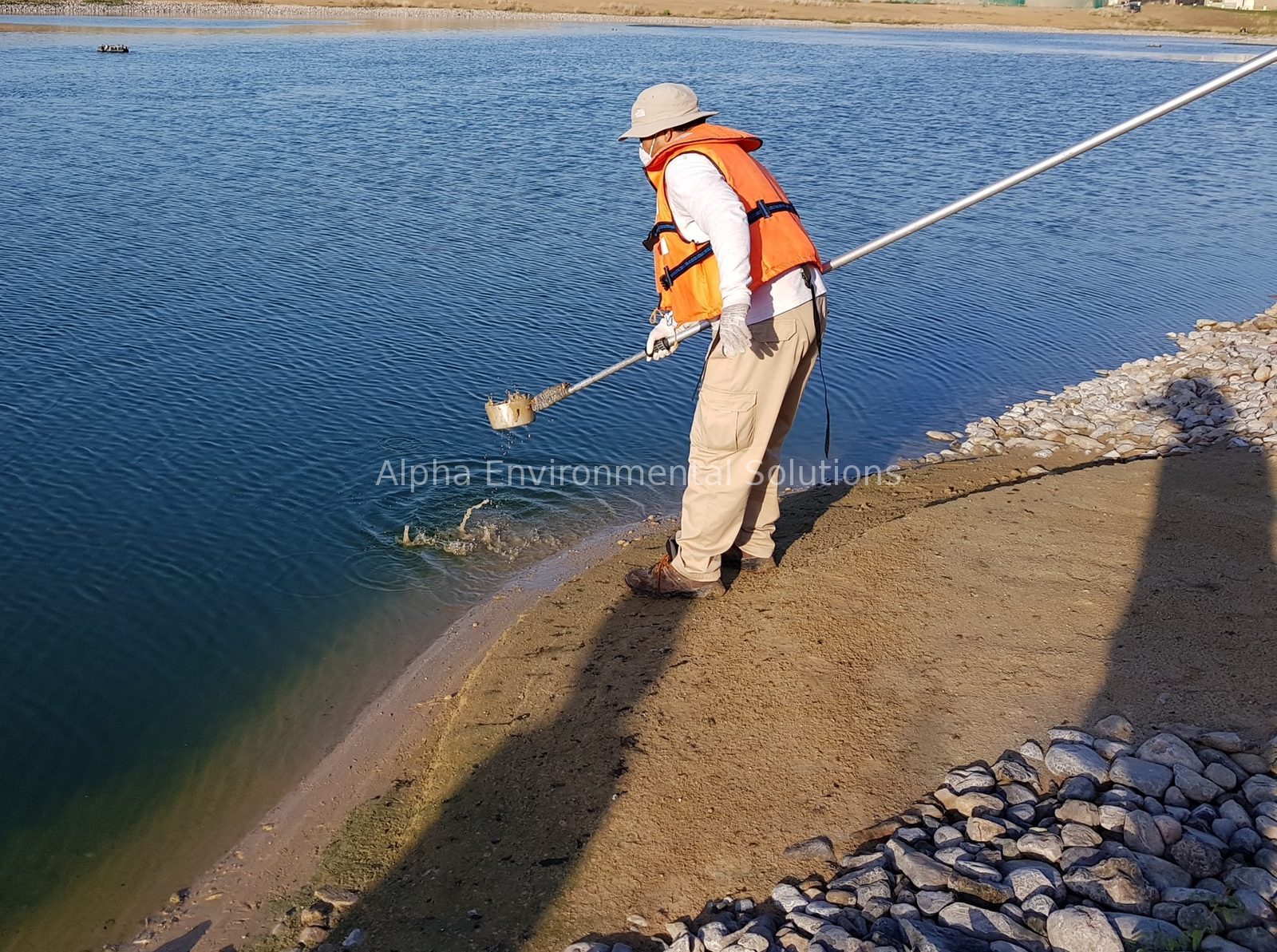 Alpha Environmental Solutions technician collecting a sample from the edge of a community lake with a long-handled sampler